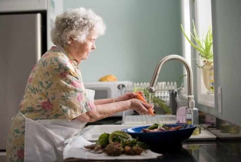 An elderly woman washing vegetables in kitchen.